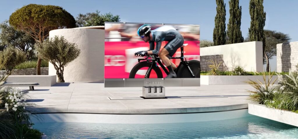 Large outdoor TV by a pool displays a cyclist competing in a race, with modern landscaping in the background.