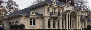 Large two-story beige house with decorative columns, arched windows, balconies, and manicured shrubs in front.