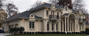 Large two-story beige house with decorative columns, arched windows, balconies, and manicured shrubs in front.