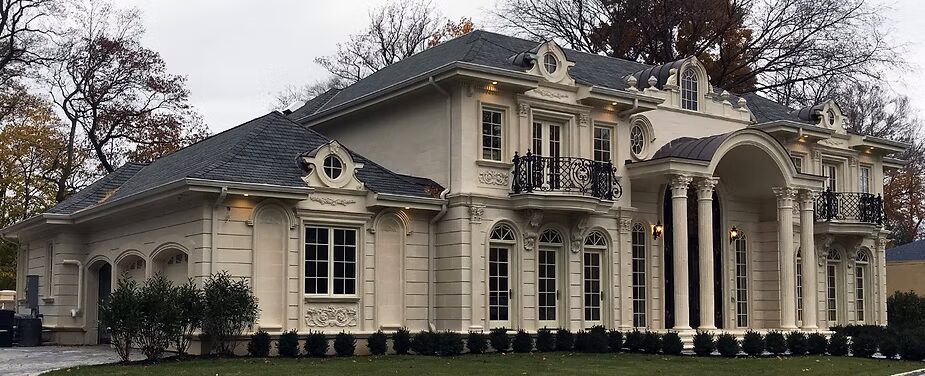 Large two-story beige house with decorative columns, arched windows, balconies, and manicured shrubs in front.