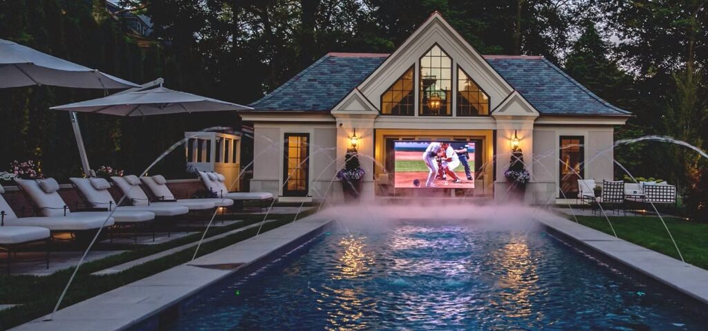 Outdoor pool with lounge chairs faces a pool house featuring a large TV showing a baseball game, with water fountains spraying.