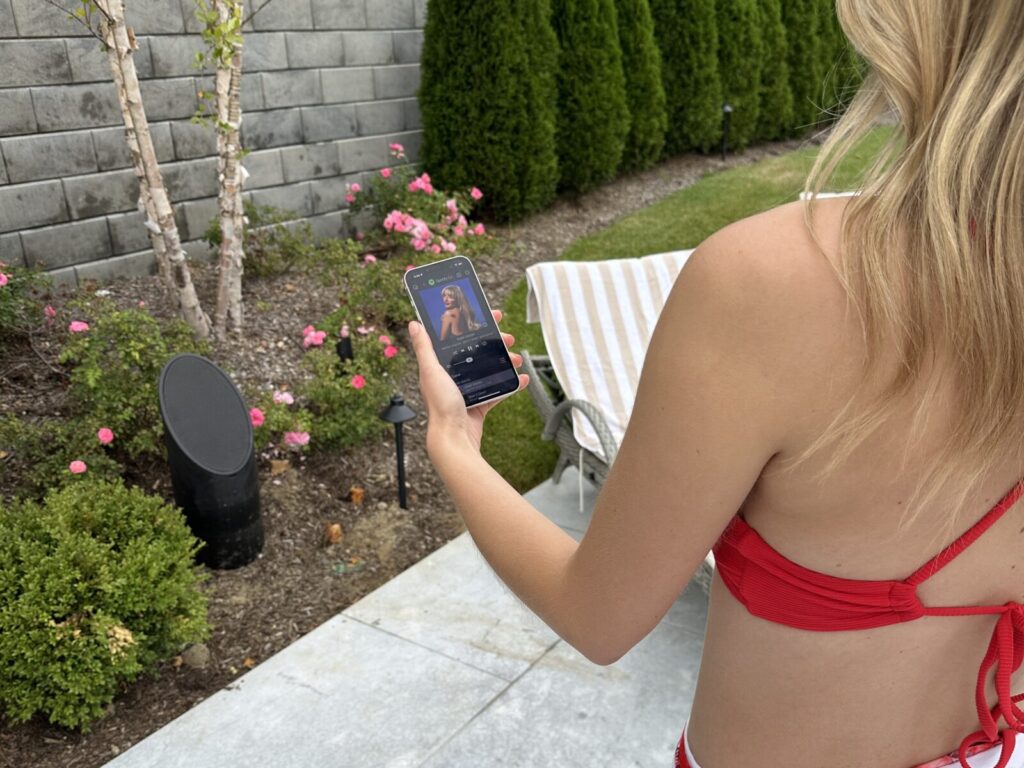 Woman in a red bikini holds a smartphone outdoors, playing music near a lounge chair and garden.