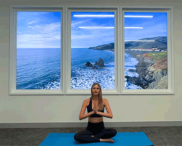 A woman practices yoga in a seated pose on a mat indoors, with large windows showing an ocean view behind her.