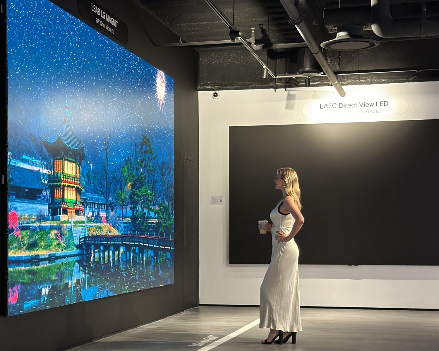 A woman in a white dress stands in front of a large, brightly lit digital display showing a scenic night view.