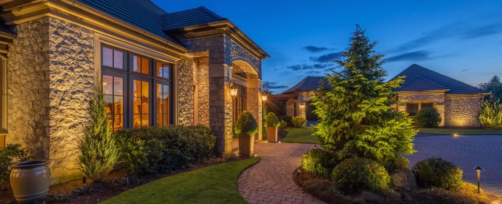 Stone house with large windows and garden, illuminated by outdoor lights at dusk with a brick path and landscaping.