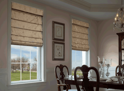 Dining room with wooden furniture, crystal decor, and two windows covered by beige Roman shades overlooking a grassy lawn.