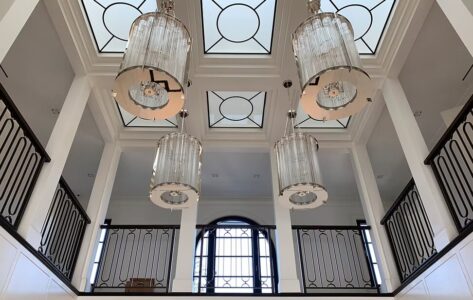 Large chandeliers hang from a high ceiling with geometric skylights above a mezzanine with black railings.