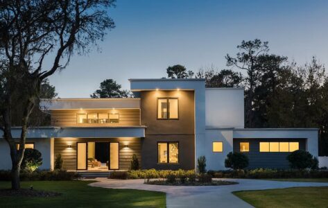 Modern two-story house with large windows, flat roof, and outdoor lighting, set against trees at dusk.