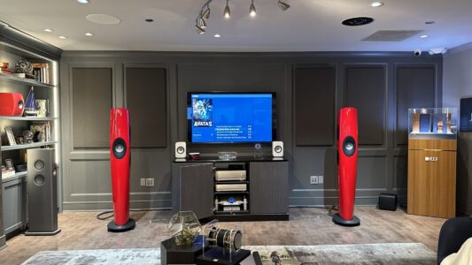 A modern home theater room with red floor speakers, a flat-screen TV, audio equipment, and grey paneled walls.