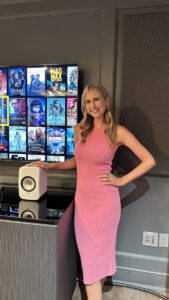 Woman in a pink dress stands indoors beside a TV displaying movie posters and a white speaker on a console.