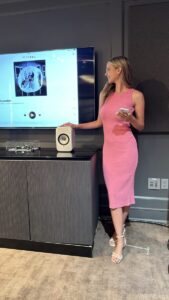 Woman in a pink dress stands beside a speaker and TV displaying a music streaming app screen, holding a smartphone.