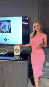 Woman in a pink dress stands beside a speaker, holding a device, with a Tidal music screen displayed on the TV behind her.