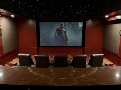 A home theater with leather seats faces a large screen displaying a scene from a Batman movie under a starry ceiling.