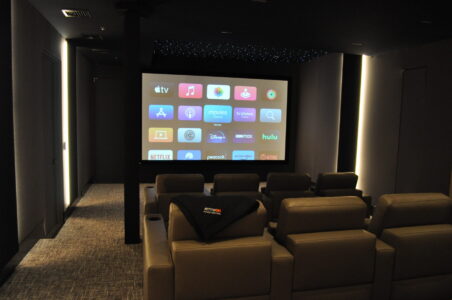 A home theater room with reclining seats faces a large screen displaying various streaming service app icons.
