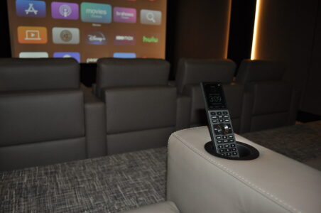 A remote control rests in a cup holder on a theater seat, with a TV screen displaying streaming apps in the background.