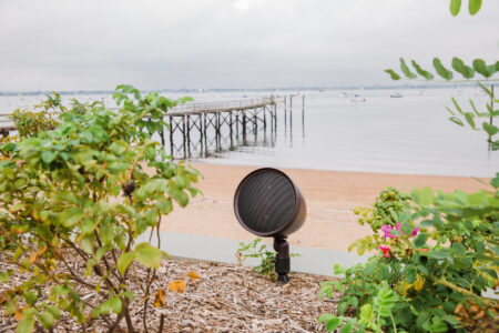 A round outdoor speaker is positioned among plants with a wooden pier and calm water in the background.