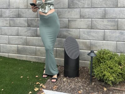 Woman in a green skirt and sandals stands outdoors by a speaker, holding a phone, with bushes and a stone wall behind her.