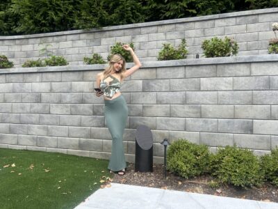 Woman in a patterned crop top and long skirt poses outdoors near a stone wall and landscaping while holding a phone.