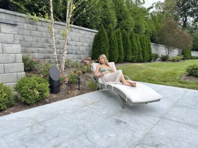 A woman relaxes on a lounge chair on a patio, surrounded by greenery and landscaped garden beds.