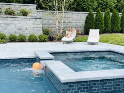 A person sits on a lounge chair beside a pool with a floating orange ball and landscaped greenery in the background.