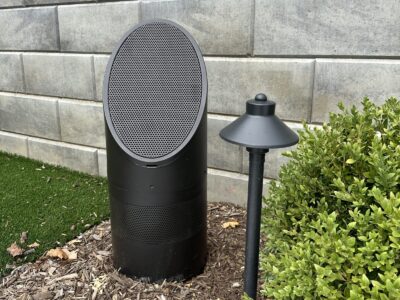 A black outdoor speaker and a path light are placed in mulch near a green shrub and a gray brick wall.