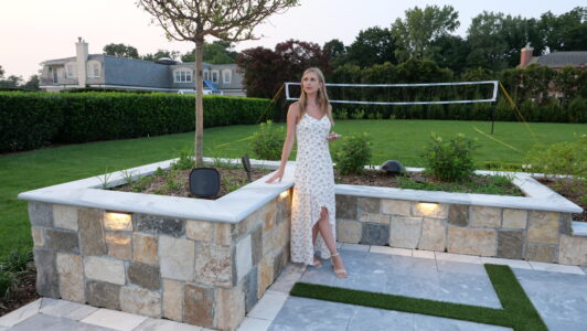 A woman in a white floral dress stands by a raised garden bed in a backyard with a volleyball net in the background.