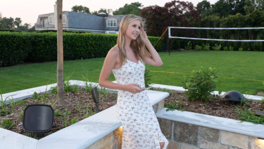 Woman in a white floral dress stands outdoors near a garden bed, holding a phone, with a volleyball net in the background.