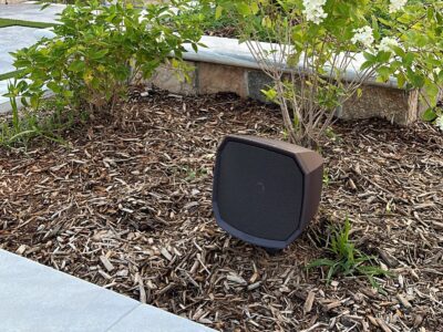 A black outdoor speaker is placed in a mulched garden bed near green plants and a stone border.