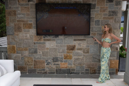A woman in a patterned outfit stands next to a large outdoor TV displaying a tennis match on a stone wall.