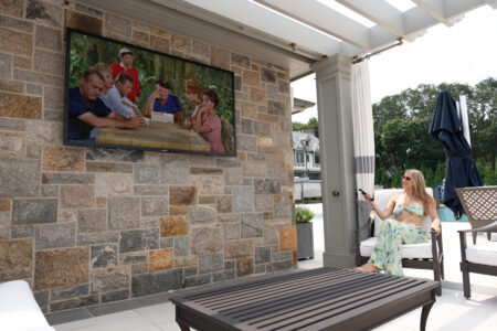 A woman sits on an outdoor patio watching a TV mounted on a stone wall, displaying a classic TV show.