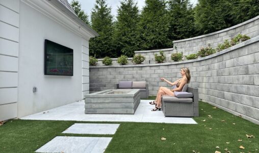 A woman sits alone on a patio sofa watching a wall-mounted TV in a modern, fenced outdoor area with artificial grass.