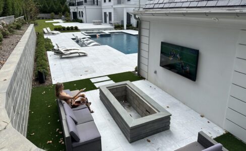 Woman sits on outdoor couch near a fire pit watching TV beside a pool in a modern backyard.