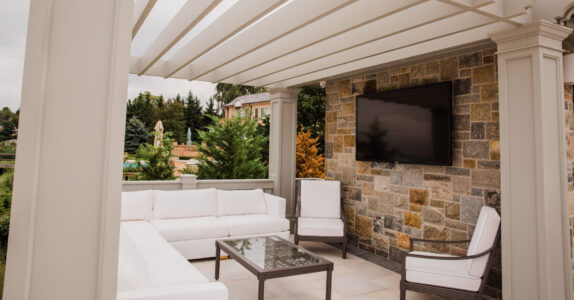 Outdoor patio with white sectional sofa, glass coffee table, armchair, TV mounted on a stone wall, and pergola overhead.