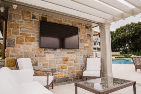 Flat-screen TV mounted on a stone wall in a patio seating area beside a pool with white cushioned chairs and a glass table.