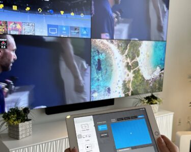 A person holds a tablet in front of a large TV displaying multiple split-screen video feeds and apps.