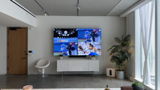 Modern living room with large TV displaying multiple sports broadcasts, white chair, plants, and natural light from tall windows.