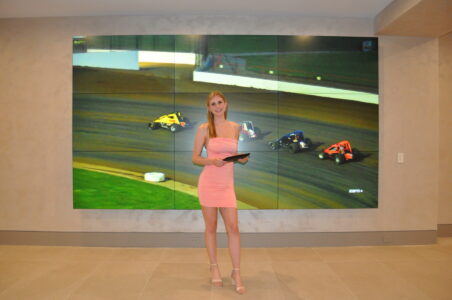 A woman in a pink dress stands indoors holding a tablet, with a race car event displayed on large screens behind her.
