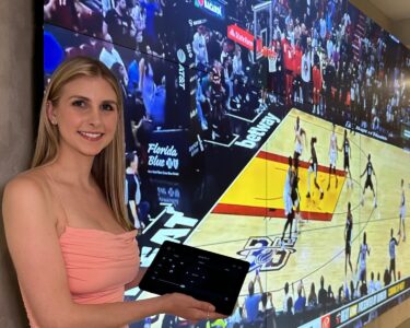 A woman holding a tablet stands in front of a large screen displaying a basketball game in progress.