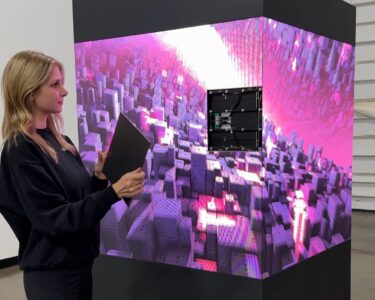 A woman replaces a panel on a large, cube-shaped digital display showing a pink and purple cityscape.