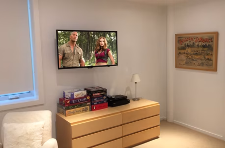 A wall-mounted TV displaying a movie above a dresser with board games, lamp, and framed poster in a tidy room.