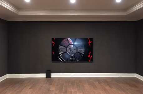 A flat-screen TV is mounted on a dark wall, showing a video game, with hardwood floors and recessed ceiling lights.