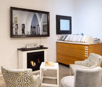 Two patterned chairs face a small table and modern fireplace in a bright room with art and a large mirror above the mantel.