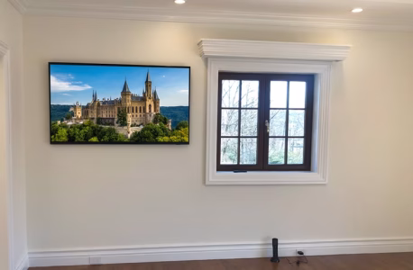 A wall-mounted TV displaying a castle scene is next to a window with a black frame in a plain room.