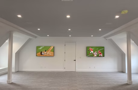 Spacious room with two mounted TVs, one showing baseball and the other football, and gray carpet flooring.