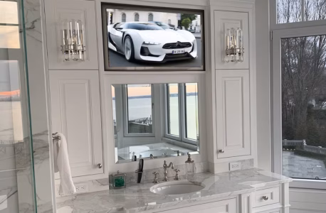 A bathroom vanity with a sink, mirror, and TV displaying a white sports car above the mirror, overlooking a waterfront view.