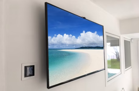 A wall-mounted flat screen TV displays a beach scene in a modern white room with windows.