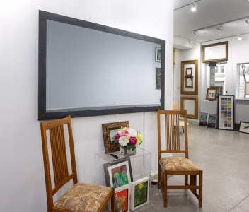 Gallery interior with two wooden chairs, a large mirror, framed artwork, flowers in a vase, and white walls.