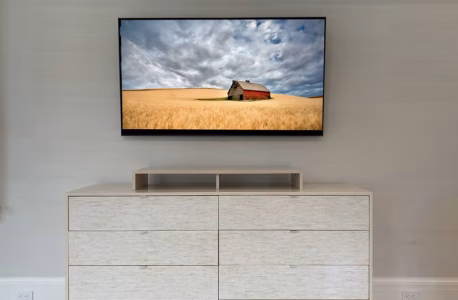 A wall-mounted TV displays a barn in a wheat field above a light-colored dresser with six drawers.