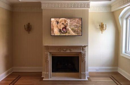 A TV above a decorative fireplace displays a roaring lion cub in an elegant, beige living room with wall sconces.