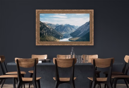 A framed landscape photograph of mountains and a river hangs on a dark wall above a dining table with wooden chairs.